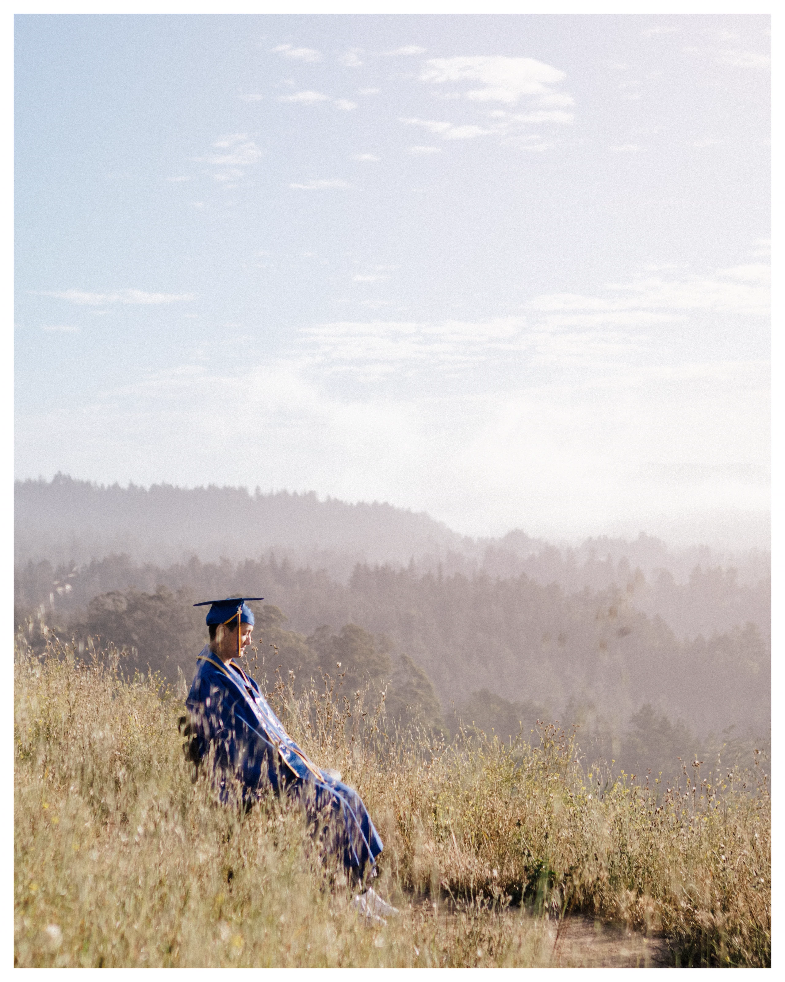 A graduate in a blue cap and gown sits in tall grass above wooded hills in Santa Cruz.