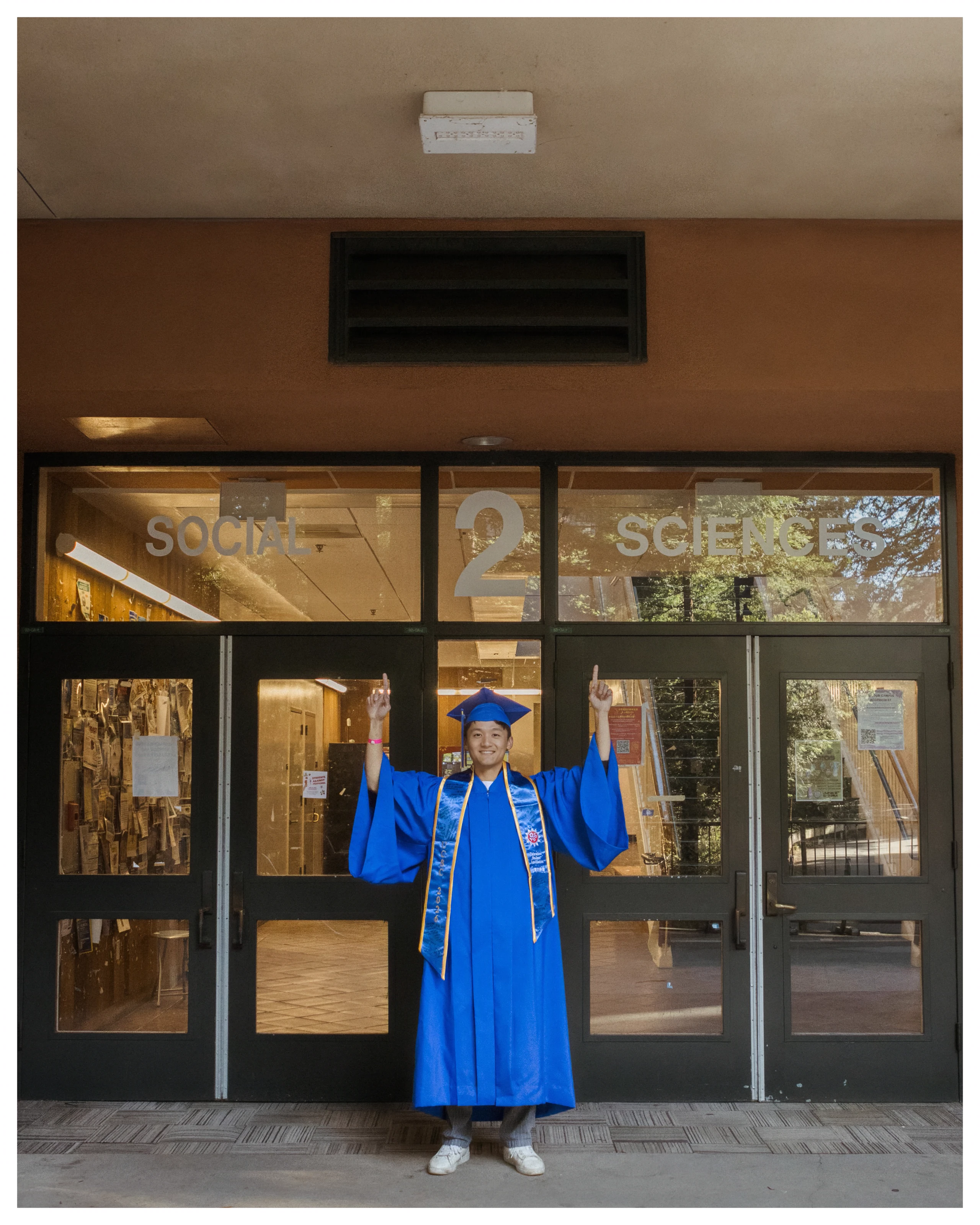 The graduate raises both hands outside the glass entrance to Social Sciences 2.